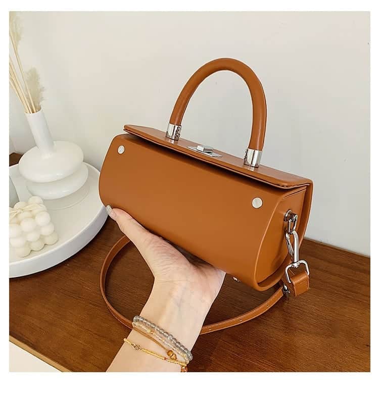 Photo d'un sac à main bandoulière cylindrique en cuir synthétique camel tenu dans une main au dessus d'u comptoir en bois