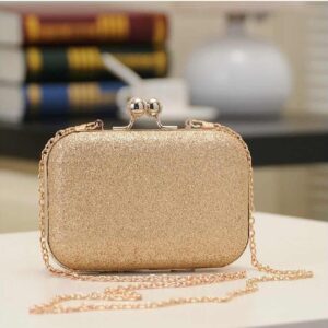 Photo d'une pochette à bandoulière doré style clutch posé sur une table avec au fond des livre et un vase Vit