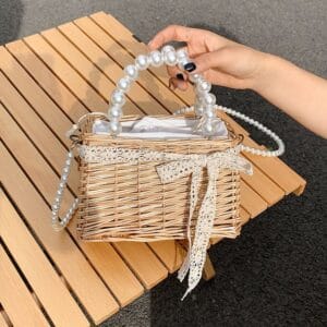 Photo d'un petit sac en osier entouré d'un ruban de dentelle avec une anse et une bandoulière en perles. Le sac est tenue par une main au dessus d'une table en bois