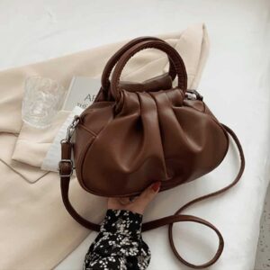 Photo d'un petit sac à main bandoulière Brun aux détails plissé tenue par une main au dessus d'une veste blazer beige.