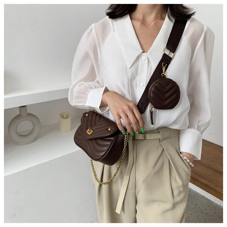 Femme portant un sac en cuir synthétique Brun en bandoulière avec une petite pochette ronde sur la sangle bandoulière. Le sac a aussi une chaine. La femme se tient debout devant un mur étagère, elle est habillée avec un pantalon beige et une chemise Vithe.