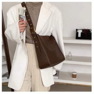 Photo d'une femme portant un sac fourre-tout bandoulière de style vintage Brun. La femme porte un pull col roulé Brun, une veste Vithe et un pantalon beige, elle se tient debout devant des étagères.
