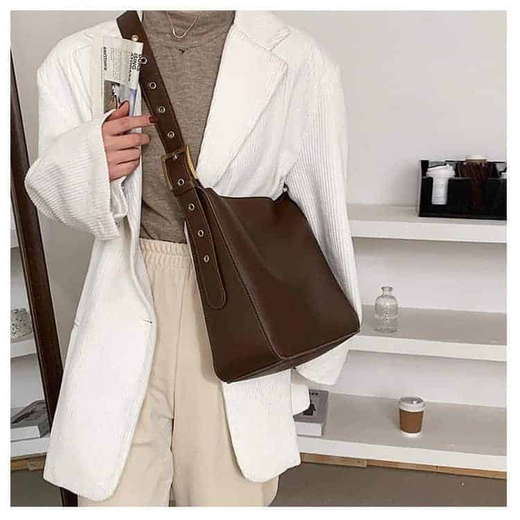 Photo d'une femme portant un sac fourre-tout bandoulière de style vintage Brun. La femme porte un pull col roulé Brun, une veste Vithe et un pantalon beige, elle se tient debout devant des étagères.