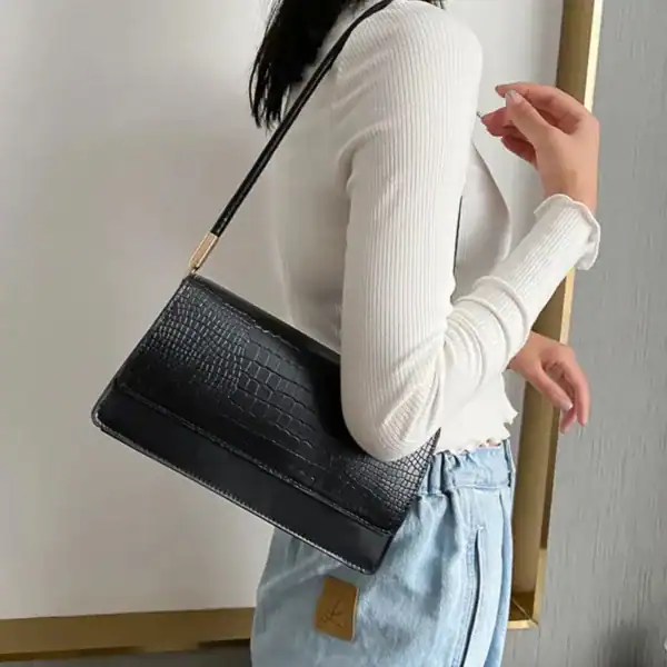 Svart krokodilsmönstrad axelväska med guld detaljer, modell på kvinna i vit tröja och jeans.