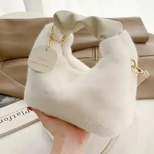 Beige pälsväska med handtag och gyllene kedja, mjuk pälsliknande yta.