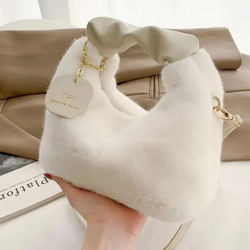 Beige pälsväska med handtag och gyllene kedja, mjuk pälsliknande yta.