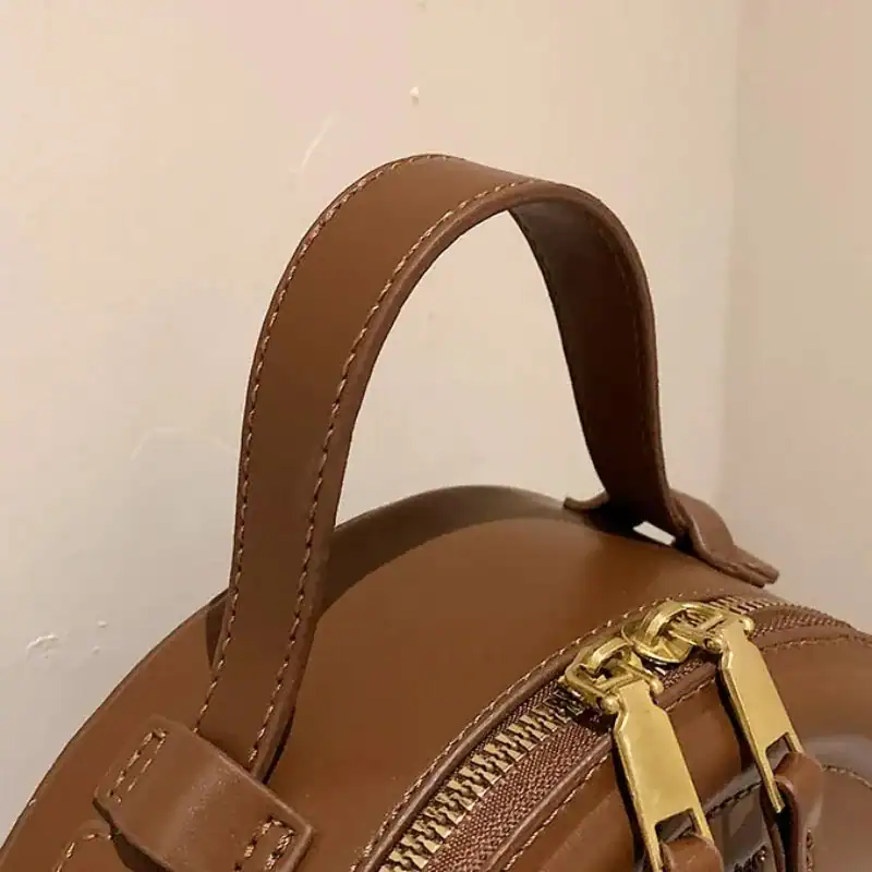 Beige rund väska med guld dragkedja och handtag.