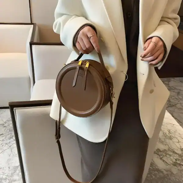 Beige rund väska med axelrem, läder, minimalistisk design.