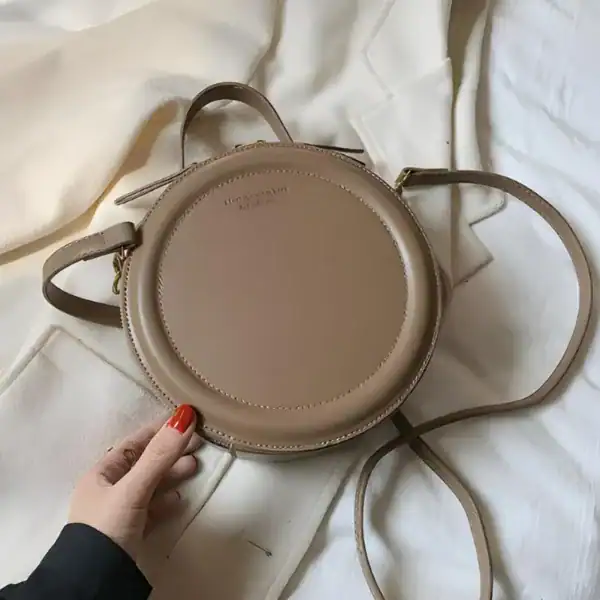 Beige rund väska med axelrem, läder, minimalistisk design.