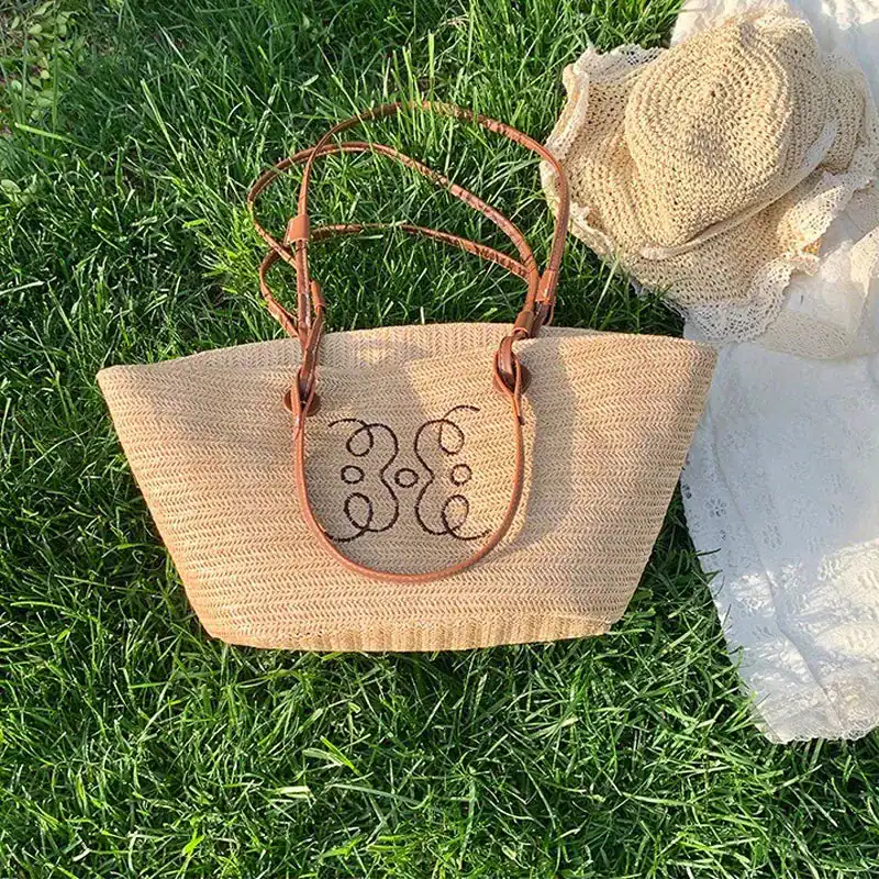 Beige stråkorg med läderhandtag och broderi, placerad på gräs.