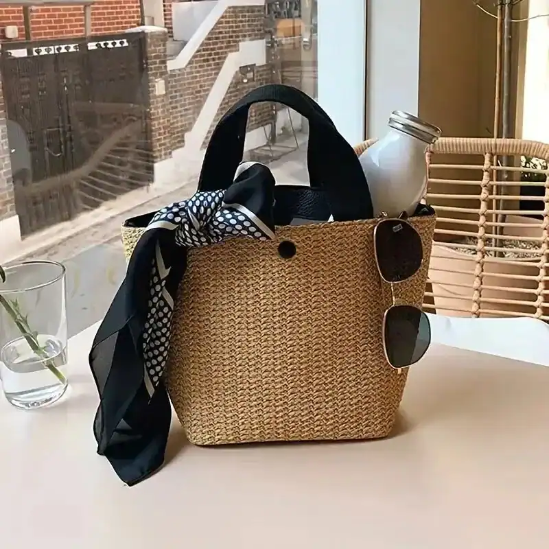 Beige stråväska med svarta handtag, solglasögon och tyg på bordet.