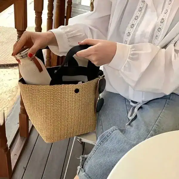 Beige stråväska med svarta handtag, hålls av kvinna i vit skjorta och jeans.