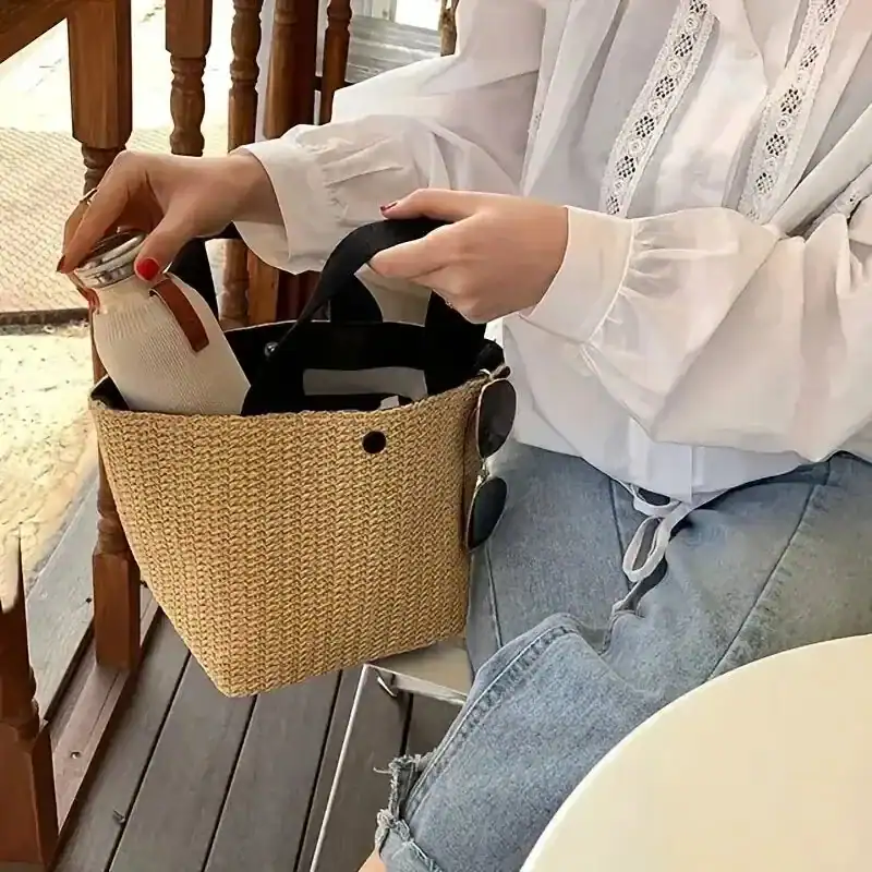 Beige stråväska med svarta handtag, hålls av kvinna i vit skjorta och jeans.
