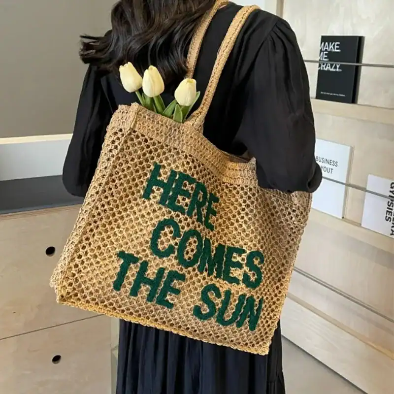 Flätad väska med text 'HERE COMES THE SUN', vita tulpaner i handtaget.