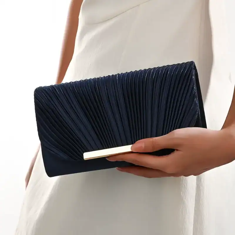 Guldig plisserad clutch i mörkblått, hålls i handen av en kvinna i vit klänning.