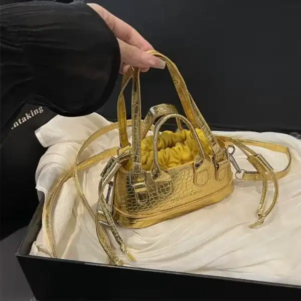 Guldväska med krokodilsmönster och gyllene detaljer, hållen i en svart aska.