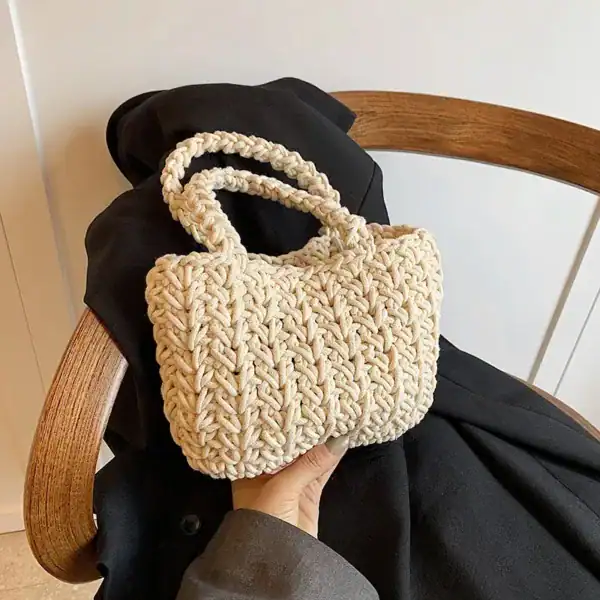 Krämvit virkad korgväska med dubbla handtag, hållen i en hand mot svart jacka och trästol.