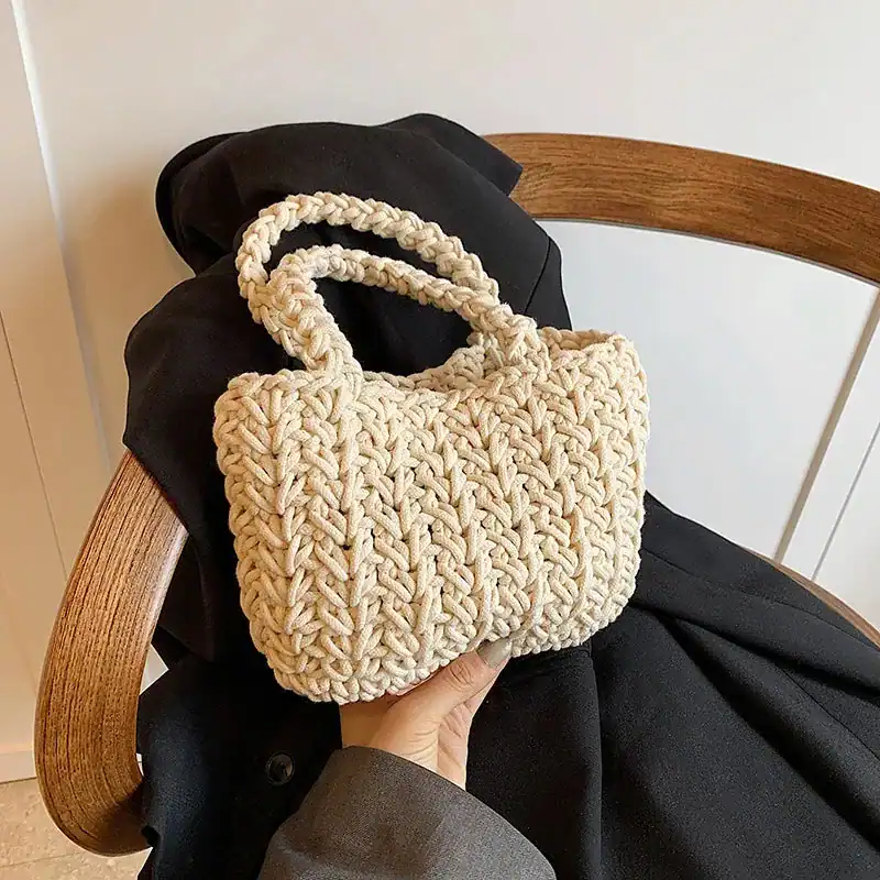 Krämvit virkad korgväska med dubbla handtag, hållen i en hand mot svart jacka och trästol.