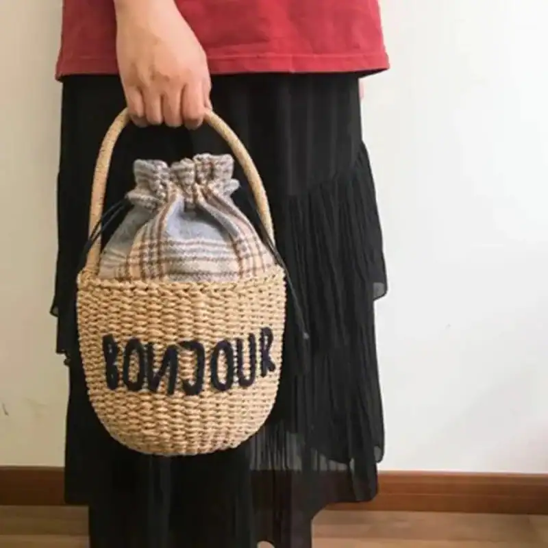 Liten stråväska med text 'BONJOUR', handtag, plädinnehåll.