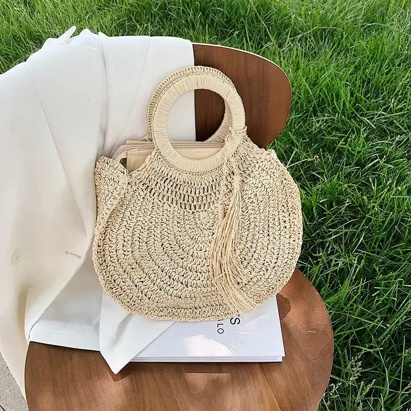 Rund halmväska med tofs, sommarväska, naturfärgad, sittande på trästol i gräs.