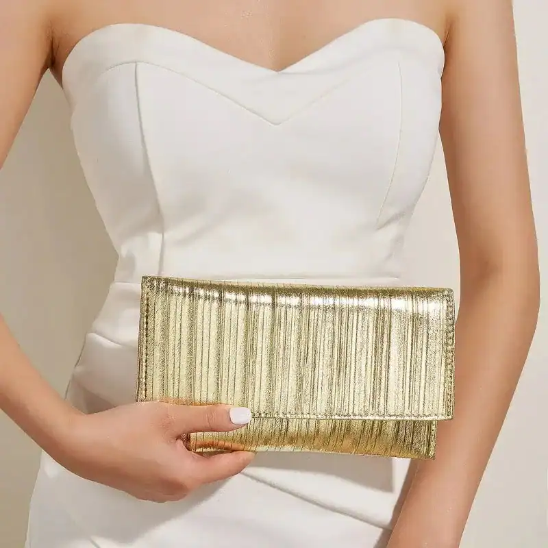 Silver plisserad clutch i guld, hållen av en kvinna i vit klänning.
