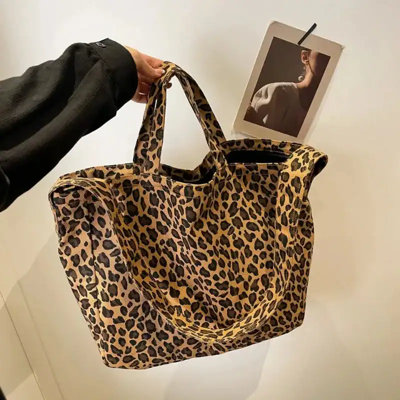 Stor leopard shoppingväska i beige och svart, hållen i handen mot vit vägg.