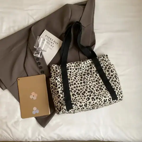 Vit leopard mönstrad axelväska med svarta handtag, placerad på säng med kläder och tablet.